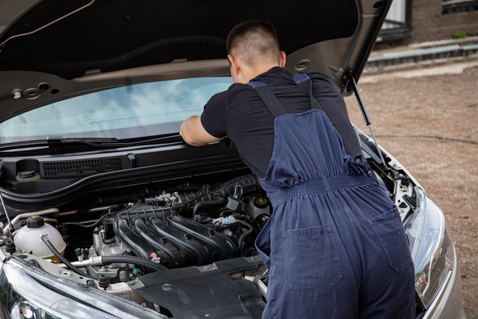A mechanic in overalls fixing a car engine outdoors on a sunny day.